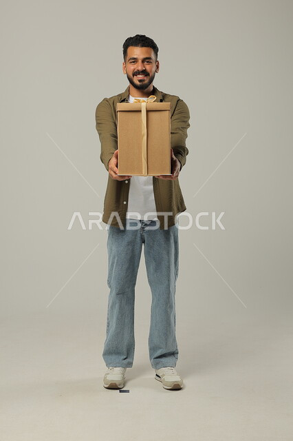 تقديم الهدية بحماس وسعادة، الاحتفال بالأعياد والمناسبات السعيدة، إعطاء الهدايا للأهل والأصدقاء المقربين, بورتريه لشاب عربي خليجي سعودي وسيم يرتدي الزي الكاجوال يحمل بيديه علبة هدايا مغلفة, الجسم بالطول الكامل، خلفية رمادية