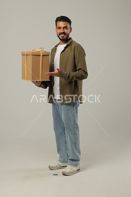 الوقوف والإشارة إلى الهدية بإيماءات السرور، الاحتفال بالأعياد والمناسبات السعيدة، تقديم الهدايا للأهل والأصدقاء المقربين, بورتريه لشاب عربي خليجي سعودي وسيم يرتدي الزي الكاجوال يحمل بيديه علبة هدايا مغلفة, الجسم بالطول الكامل، خلفية رمادية