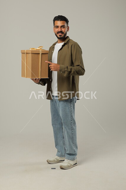 الإشارة إلى الهدية بإيماءات السرور، الاحتفال بالأعياد والمناسبات السعيدة، تقديم الهدايا للأهل والأصدقاء المقربين, بورتريه لشاب عربي خليجي سعودي وسيم يرتدي الزي الكاجوال يحمل بيديه علبة هدايا مغلفة, الجسم بالطول الكامل، خلفية رمادية
