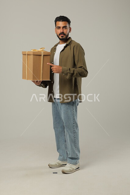 النظر إلى الكاميرا والإشارة إلى الهدية، الاحتفال بالأعياد والمناسبات السعيدة، تقديم الهدايا للأهل والأصدقاء المقربين, بورتريه لشاب عربي خليجي سعودي وسيم يرتدي الزي الكاجوال يحمل بيديه علبة هدايا مغلفة, الجسم بالطول الكامل، خلفية رمادية