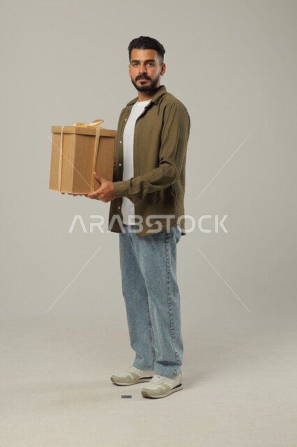 التعبير عن المحبة بتقديم الهدايا, مفهوم الإهداء والتقدير، أعياد ومفاجآت ومناسبات سعيدة, بورتريه لشاب عربي خليجي سعودي وسيم يرتدي الزي الكاجوال يحمل بيديه علبة هدايا مغلفة, الجسم بالطول الكامل، خلفية رمادية