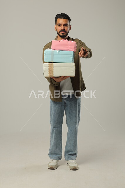  عدد من العلب المغلفة والمرتبة فوق بعضها البعض، المباركات والأفراح وأعياد الميلاد، تقديم الهدية بالمناسبات العائلية السعيدة, بورتريه لشاب عربي خليجي سعودي وسيم يرتدي الزي الكاجوال يحمل بيديه الهدايا الملونة, الجسم بالطول الكامل، خلفية رمادية