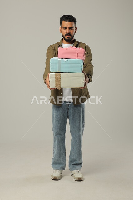 النظر إلى الهدايا باستغراب ودهشة، أعياد الميلاد والمباركات والأفراح، تقديم الهدية بالمناسبات العائلية السعيدة, بورتريه لشاب عربي خليجي سعودي وسيم يرتدي الزي الكاجوال يحمل بيديه عدد من العلب المغلفة والمرتبة فوق بعضها البعض, الجسم بالطول الكامل، خلفية رمادية