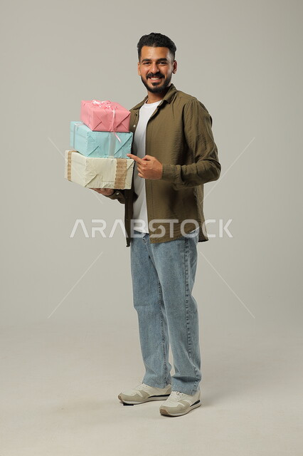 Pointing to gifts with gestures of happiness, birthdays, blessings and joys, giving a gift on happy family occasions, portrait of a handsome Saudi Gulf Arab young man wearing a casual outfit holding in his hands a number of wrapped boxes arranged on top of each other, full length body, gray background