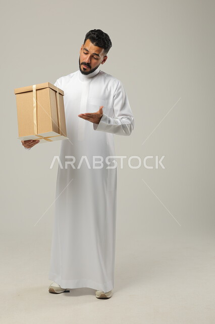 النظر للهدية بإيماءات البشاشة، زيادة روابط المحبة بالإهداء, أعياد الميلاد والمباركات والأفراح, تقديم الهدايا بالمناسبات العائلية السعيدة,بورتريه لشاب عربي خليجي سعودي وسيم يرتدي الثوب التقليدي يمسك بيده علبة ملفوفة بشريط أنيق, الجسم بالطول الكامل, خلفية رمادية