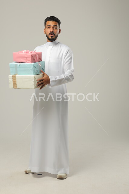 الاحتفال بالأعياد السعيدة, المباركات والتهنئات بالنجاح والأفراح, تقديم الهدية في مناسبات الأهل والأصدقاء المقربة, بورتريه لرجل عربي خليجي سعودي مبتسم يرتدي الثوب التقليدي يحمل بيديه علب هدايا مغلفة بألوان أنيقة, الجسم بالطول الكامل, خلفية رمادية