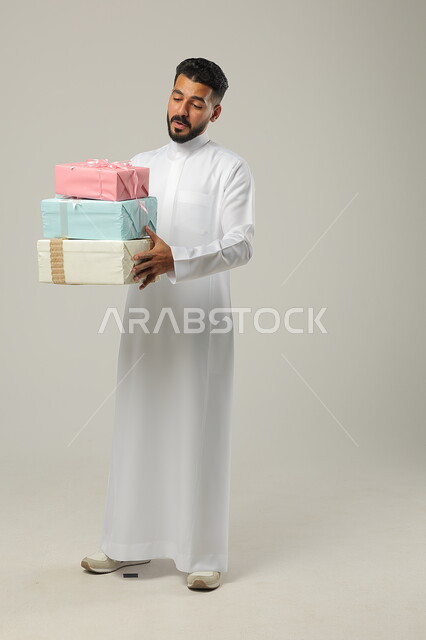 المباركات والتهنئات بالنجاح والأفراح, الاحتفال بالأعياد السعيدة, تقديم الهدية في مناسبات الأهل والأصدقاء المقربة, بورتريه لرجل عربي خليجي سعودي مبتسم يرتدي الثوب التقليدي يحمل بيديه علب هدايا مغلفة بألوان أنيقة, الجسم بالطول الكامل, خلفية رمادية