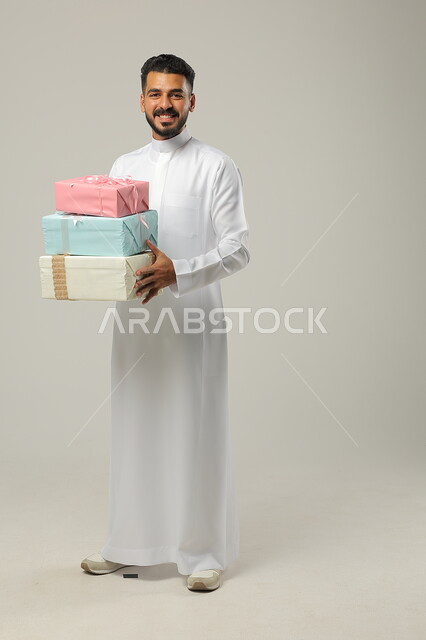 التهنئات والمباركات بالنجاح والأفراح, الاحتفال بالأعياد السعيدة, تقديم الهدية في مناسبات الأهل والأصدقاء المقربة, بورتريه لرجل عربي خليجي سعودي مبتسم يرتدي الثوب التقليدي يحمل بيديه علب هدايا مغلفة بألوان أنيقة, الجسم بالطول الكامل, خلفية رمادية