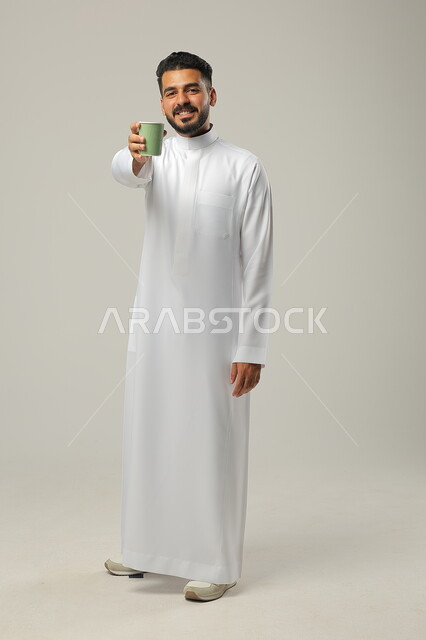Providing authentic Arab hospitality, sipping a delicious hot drink, looking at the camera and enjoying a favorite drink, portrait of a Saudi Gulf Arab young man wearing a traditional dress and holding a cup of coffee in his hand, luxurious Arabic coffee, enjoying drinks in cafes and ready-to-eat restaurants, full-length body, gray background