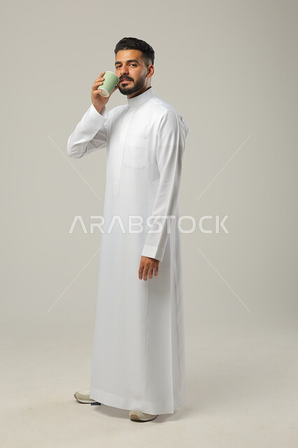 النظر للكاميرا والاستمتاع بتناول المشروبات المفضلة, احتساء مشروب ساخن لذيذ, بورتريه لشاب عربي خليجي سعودي يرتدي الثوب التقليدي ويحمل بيده كوب من القهوة, البن العربي الفاخر الأصيل, التلذذ بمشاريب الكافيهات والمطاعم الجاهزة، الجسم بالطول الكامل، خلفية رمادية