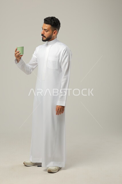 Enjoying drinking favorite drinks, sipping a delicious hot drink, portrait of a Saudi Gulf Arab young man wearing a traditional dress and holding a cup of coffee in his hand, authentic fine Arabic coffee, enjoying drinks in cafes and ready-to-eat restaurants, full-length body, gray background
