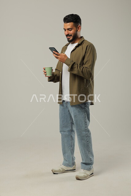 متابعة آخر المستجدات والأخبار باستخدام الهاتف المحمول, التلذذ باحتساء مشاريب الكافيهات والمطاعم الجاهزة، الاستمتاع بتناول المشروبات المفضلة, بورتريه لشاب عربي خليجي سعودي يرتدي الزي الكاجوال يحمل بيده كوب قهوة، الجسم بالطول الكامل, خلفية رمادية
