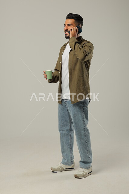 Communicating with family and friends via mobile phone, drinking a hot and delicious drink, enjoying drinks in cafes and ready-to-eat restaurants, enjoying favorite drinks, portrait of a Saudi Gulf Arab young man wearing a casual outfit, holding a cup of coffee in his hand, body full length, gray background