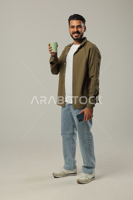 النظر للكاميرا بإيماءات السرور, احتساء مشروب ساخن ولذيذ, التلذذ بمشاريب الكافيهات والمطاعم الجاهزة، الاستمتاع بتناول المشروبات المفضلة, بورتريه لشاب عربي خليجي سعودي يرتدي الزي الكاجوال يحمل بيده كوب قهوة، الجسم بالطول الكامل, خلفية رمادية