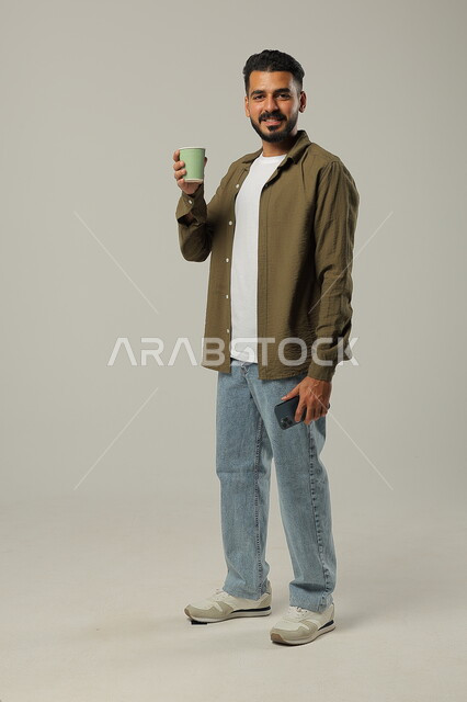  احتساء مشروب ساخن ولذيذ, التلذذ بمشاريب الكافيهات والمطاعم الجاهزة، الاستمتاع بتناول المشروبات المفضلة, بورتريه لشاب عربي خليجي سعودي يرتدي الزي الكاجوال يحمل بيده كوب قهوة وينظر للكاميرا بإيماءات السرور, الجسم بالطول الكامل, خلفية رمادية