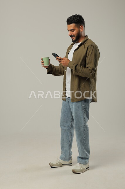 تصفح مواقع التواصل الاجتماعي باستخدام الهاتف المحمول, التلذذ بمشاريب الكافيهات والمطاعم الجاهزة، الاستمتاع بتناول المشروبات المفضلة, احتساء مشروب ساخن لذيذ, بورتريه لشاب عربي خليجي سعودي يرتدي الزي الكاجوال يحمل بيده كوب قهوة, الجسم بالطول الكامل, خلفية رمادية