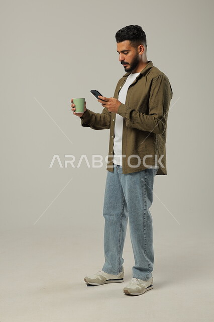 التلذذ بمشاريب الكافيهات والمطاعم الجاهزة، الاستمتاع بتناول المشروبات المفضلة, احتساء مشروب ساخن لذيذ, بورتريه لشاب عربي خليجي سعودي يرتدي الزي الكاجوال يمسك بيده الهاتف المحمول, تصفح مواقع التواصل الاجتماعي, الجسم بالطول الكامل, خلفية رمادية
