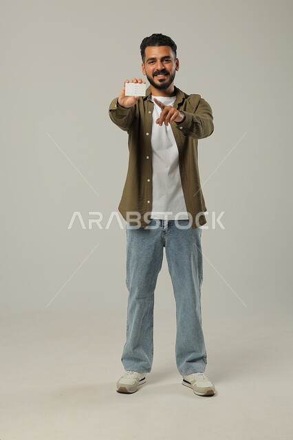 الإشارة إلى البطاقة التوضيحية بإيماءات السعادة, الوقوف باستقامة وثقة، النظر إلى الكاميرا بإيماءات السرور و الرضا، بورتريه لرجل عربي خليجي سعودي مبتسم يرتدي الزي الكاجوال يحمل بيده كرت تعريفي, الجسم بالطول الكامل، خلفية رمادية