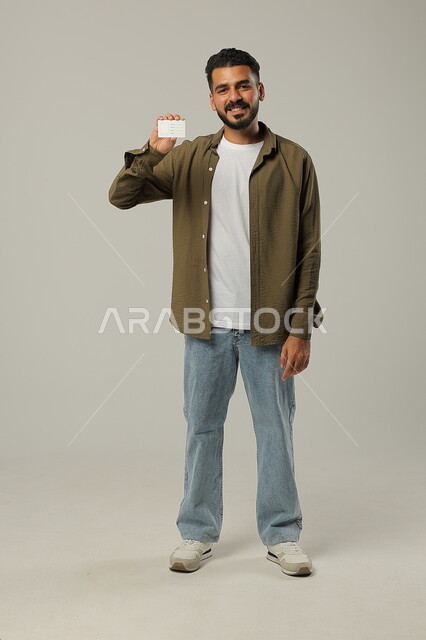 النظر الى الكاميرا بإيماءات السرور و الرضا، بورتريه لرجل عربي خليجي سعودي مبتسم يرتدي الزي الكاجوال و يحمل بيده بطاقة تعريفية, الوقوف باستقامة وثقة، استخدام بطاقات توضيحية, الجسم بالطول الكامل، خلفية رمادية