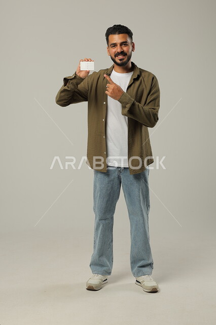 الوقوف باستقامة وثقة، النظر إلى الكاميرا بإيماءات السرور و الرضا، بورتريه لرجل عربي خليجي سعودي مبتسم يرتدي الزي الكاجوال و يحمل بيده بطاقة تعريفية ويشير إليها, استخدام بطاقات توضيحية, الجسم بالطول الكامل، خلفية رمادية