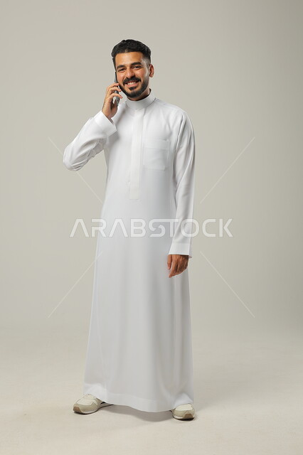تلقي الأخبار السارة، استخدام التقنيات الحديثة والتكنولوجيا، بورتريه لشاب عربي سعودي خليجي يرتدي الثوب التقليدي، النظر الى الكاميرا بايماءات السعادة والسرور، التواصل مع الاهل والاقارب عبر الهاتف المحمول، الجسم الطول الكامل خلفية رمادية