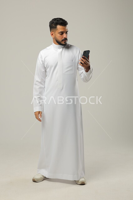 Making a video call, using modern techniques and technology, live broadcast over the Internet, portrait of a young Saudi Arab Gulf man wearing a traditional dress, communicating with family and relatives via mobile phone, full-length body, gray background
