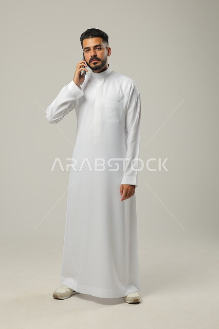 استخدام التقنيات الحديثة والتكنولوجيا، بورتريه لشاب عربي سعودي خليجي يرتدي الثوب التقليدي يتواصل مع الاهل والاقارب عبر الهاتف المحمول، الجسم بالطول الكامل، خلفية رمادية