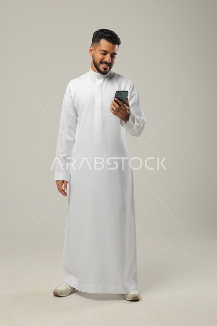 اجراء مكالمة فيديو مع الاصدقاء، بث مباشر عبر الانترنت، استخدام التقنيات الحديثة والتكنولوجيا، بورتريه لشاب عربي سعودي خليجي يرتدي الثوب التقليدي ويتواصل مع الاهل والاقارب عبر الهاتف المحمول، ايماءات السعادة والسرور، الجسم بالطول الكامل، خلفية رمادية