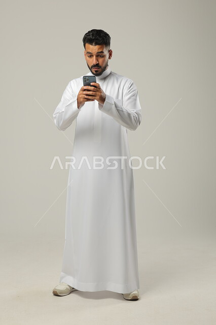 تلقي الاخبار المفاجئة، النظر الى الهاتف المحمول بايماءات الدهشة، استخدام التقنيات الحديثة والتكنولوجيا، بورتريه لشاب عربي سعودي خليجي يرتدي الثوب التقليدي، التواصل مع الاهل والاقارب، الجسم بالطول الكامل، خلفية رمادية