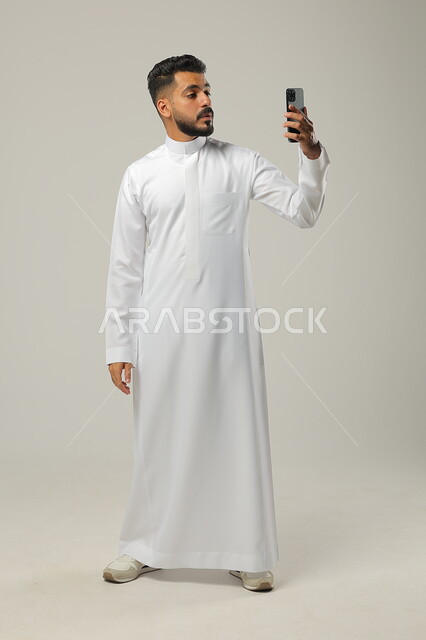 اجراء مكالمة فيديو مع الاهل والاقارب، بث مباشر عبر الانترنت، استخدام التقنيات الحديثة والتكنولوجيا، بورتريه لشاب عربي سعودي خليجي يرتدي الثوب التقليدي، التواصل مع الاصدقاء وشركاء العمل عبر الهاتف المحمول، الجسم بالطول الكامل، خلفية رمادية