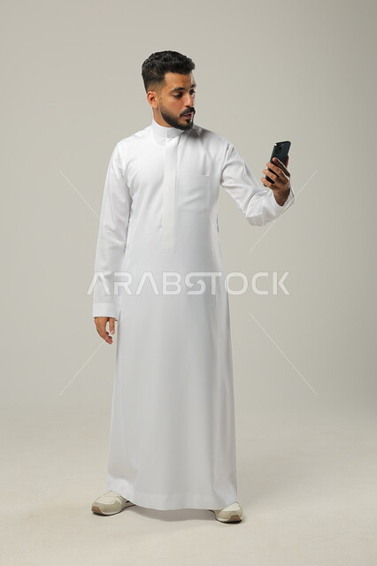 Live broadcast over the Internet, making a video call with family and relatives, using modern techniques and technology, a portrait of a young Saudi Arab Gulf man wearing a traditional dress, communicating with friends and business partners via mobile phone, full-length body, gray background