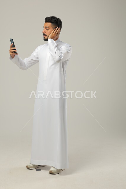 مفهوم الاستماع والاصغاء، استخدام تطبيقات الاغاني والتسجيلات الصوتية، التقنيات والتكنولوجيا الحديثة، بورتريه لشاب عربي سعودي خليجي يرتدي الثوب التقليدي يضع سماعات الهيدفون ، التواصل مع الاهل والاقارب عبر الهاتف المحمول، الجسم بالطول الكامل، خلفية رمادية