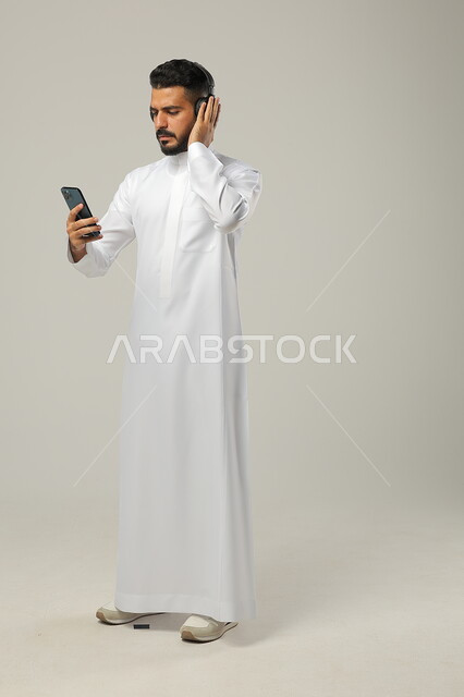 الدهشة عند سماع خبر سيء عبر الهاتف المحمول، استخدام تطبيقات الاغاني والتسجيلات الصوتية، التقنيات والتكنولوجيا الحديثة، بورتريه لشاب عربي سعودي خليجي يرتدي الثوب التقليدي يضع سماعات الهيدفون ،  مفهوم الاستماع والاصغاء، الجسم بالطول الكامل، خلفية رمادية