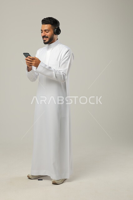 ايماءات السعادة والسرور، استخدام تطبيقات الاغاني والتسجيلات الصوتية، التقنيات والتكنولوجيا الحديثة، بورتريه لشاب عربي سعودي خليجي يرتدي الثوب التقليدي يضع سماعات الهيدفون ، التواصل مع الاهل والاقارب عبر الهاتف المحمول، مفهوم الاستماع والاصغاء، الجسم بالطول الكامل، خلفية رمادية