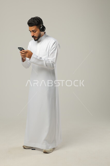   نظرات الدهشة والاستغراب، استخدام تطبيقات الاغاني والتسجيلات الصوتية، التقنيات والتكنولوجيا الحديثة، بورتريه لشاب عربي سعودي خليجي يرتدي الثوب التقليدي يضع سماعات الهيدفون ، التواصل مع الاهل والاقارب عبر الهاتف المحمول، مفهوم الاستماع والاصغاء، الجسم بالطول الكامل، خلفية رمادية