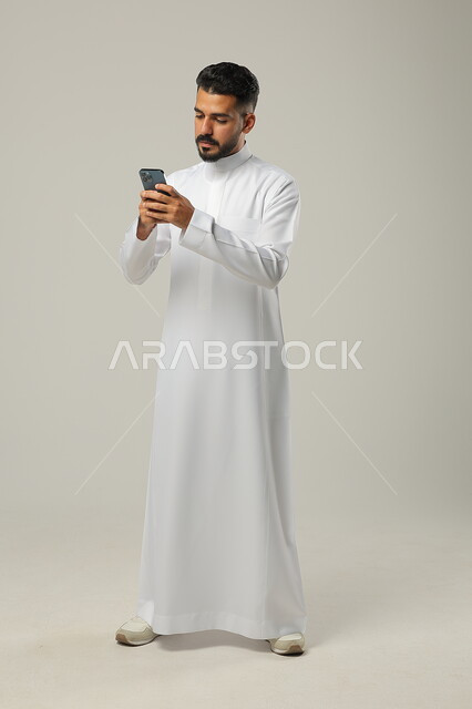 Browsing social media applications and websites, using modern techniques and technology, a portrait of a Saudi Gulf Arab young man wearing a traditional dress, talking with family and relatives via mobile phone, full-length body, gray background