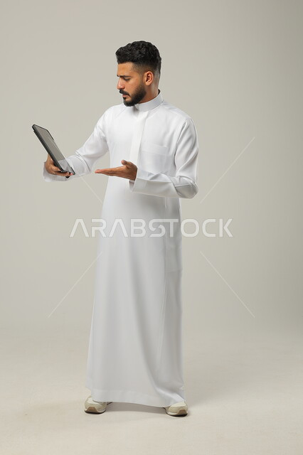 Pointing to the tablet device with movements of astonishment at something, gestures of emotion and engagement while watching something on the tablet device, a portrait of a Saudi Gulf Arab young man wearing a traditional dress making a video call and hearing surprising news, full-length body photo, gray background