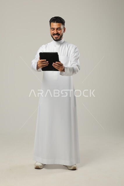  تعابير الحماس والسرور، استخدام التكنولوجيا و التقنيات الحديثة , بورتريه لرجل عربي خليجي سعودي يرتدي الثوب التقليدي يحمل بيده جهاز التابليت ،الدردشة مع الأهل والأصدقاء , تصفح برامج التواصل الاجتماعي ، تطبيقات وبرامج الأجهزة اللوحية، صورة الجسم بالطول الكامل , خلفية رمادية .