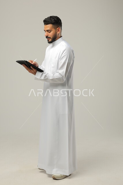 Following the latest social media news, using technology and modern techniques, a portrait of a Saudi Gulf Arab man wearing a traditional dress, holding a tablet in his hand, chatting with family and friends, browsing social media programs, full-length body photo, gray background.