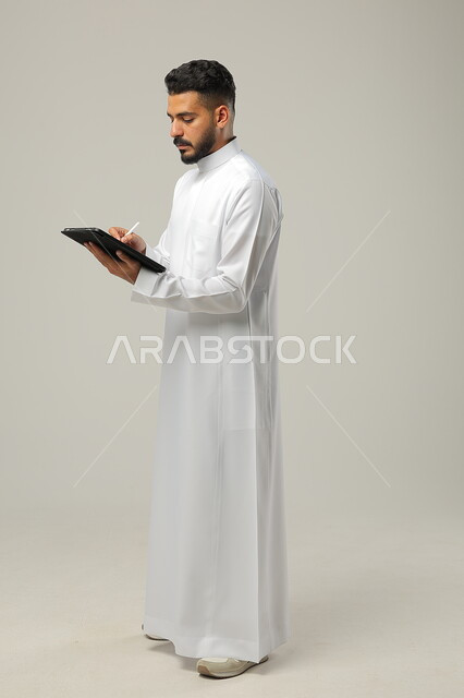 Chatting with family and friends, using technology and modern techniques, a portrait of a Saudi Gulf Arab man wearing a traditional dress holding a tablet in his hand, browsing social media programs, tablet applications and programs, full-length body photo, gray background.