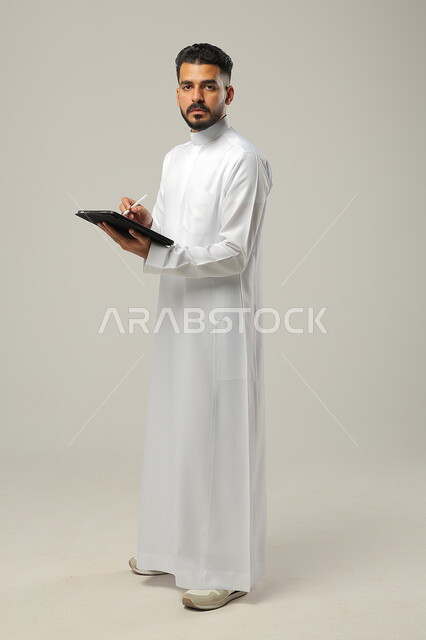 استخدام الأجهزة اللوحية الحديثة، بورتريه لرجل عربي خليجي سعودي يرتدي الثوب التقليدي يحمل بيده جهاز التابليت، تطور التقنيات والتكنولوجيا، الدردشة مع الأهل والأصدقاء، تصفح برامج التواصل الاجتماعي، صورة الجسم بالطول الكامل , خلفية رمادية .