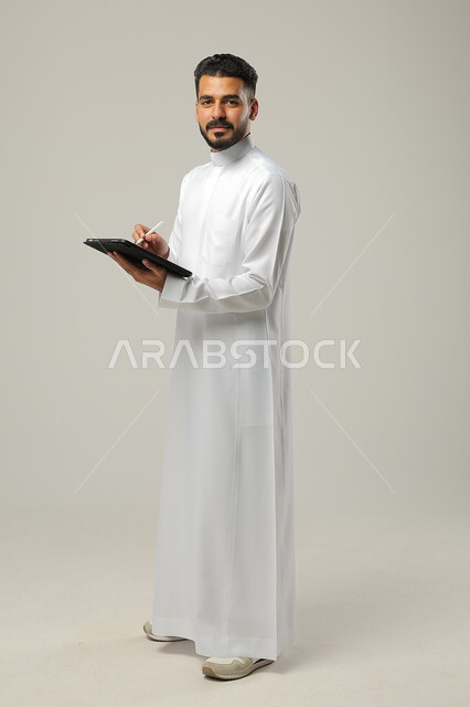 Tablet applications and programs, use of technology and modern techniques, portrait of a Saudi Gulf Arab man wearing a traditional dress, holding a tablet in his hand, chatting with family and friends, browsing social media programs, full-length body photo, gray background.