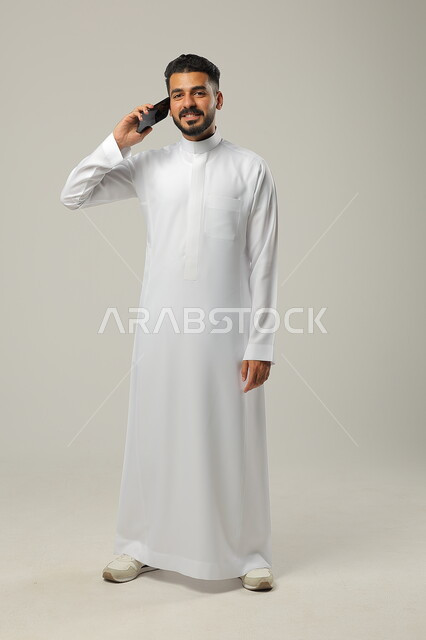 التواصل والتحدث مع العائلة والاقارب، بورتريه لرجل عربي خليجي سعودي يحمل بيده هاتف محمول، اجراء مكالمة هاتفية لمناقشة شيء ما، استخدام التقنيات والتكنولوجيا الحديثة والمتطورة، صورة الجسم كامل، خلفية رمادية