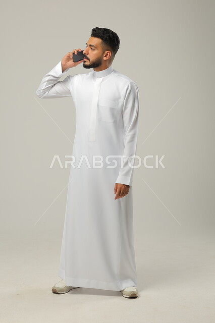 الانسجام بالمكالمة الصوتية، التواصل والتحدث مع العائلة والاقارب، بورتريه لرجل عربي خليجي سعودي يحمل بيده الهاتف المحمول يقوم باجراء مكالمات هاتفية لمناقشة شيء ما، استخدام التقنيات والتكنولوجيا الحديثة والمتطورة، صورة الجسم بالطول كامل، خلفية رمادية