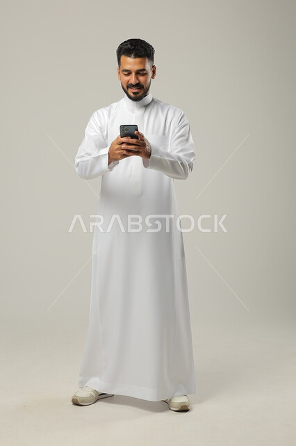 ايماءات السعادة والسرور، التواصل والتحدث مع العائلة والاقارب، بورتريه لرجل عربي خليجي سعودي يحمل بيده هاتف محمول، الدردشة مع الاصدقاء بتعابير الضحك، استخدام التقنيات والتكنولوجيا الحديثة والمتطورة، صورة الجسم كامل، خلفية رمادية