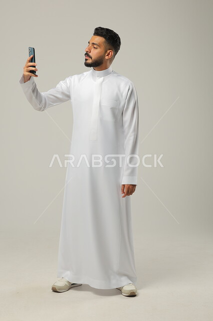 اجراء بث مباشر عبر الانترنت، التواصل والتحدث مع العائلة والاقارب، بورتريه لرجل عربي خليجي سعودي يحمل بيده الهاتف المحمول يقوم باجراء مكالمة فيديو لمناقشة شيء ما، استخدام التقنيات والتكنولوجيا الحديثة والمتطورة، صورة الجسم كامل، خلفية رمادية