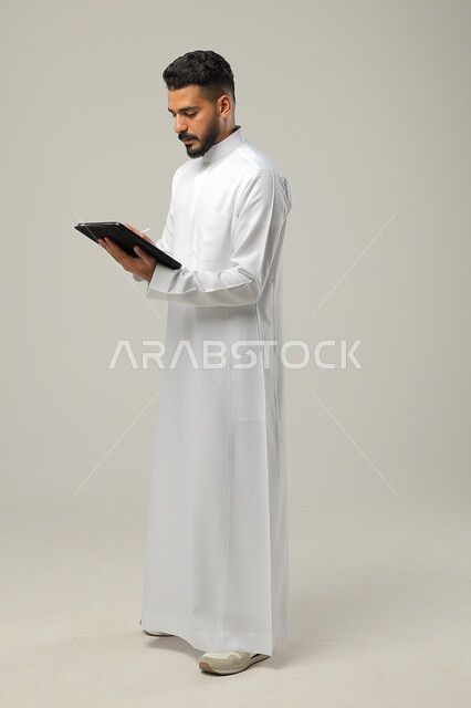 استخدام التكنولوجيا و التقنيات الحديثة , بورتريه لرجل عربي خليجي سعودي يرتدي الثوب التقليدي يحمل بيده جهاز التابليت ،الدردشة مع الأهل والأصدقاء, تطبيقات وبرامج الأجهزة اللوحية، صورة الجسم بالطول الكامل , خلفية رمادية .