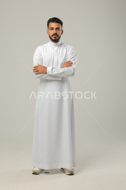 تعابير الجدية والثقة بالنفس، مفهوم الاهتمام بالمظهر الخارجي والاناقة، بورتريه لرجل عربي خليجي سعودي يرتدي الثوب الأبيض يقف باستقامة بحركات أيدي متقاطعة، النظر الى الكاميرا بايماءات العبوس، صورة الجسم بالطول الكامل، خلفية رمادية
