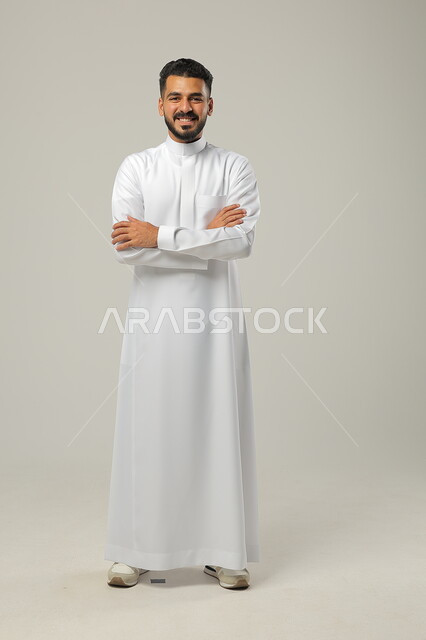 مفهوم الاهتمام في المظهر الخارجي والأناقة، بورتريه لرجل خليجي عربي سعودي مبتسم  يقف بتعابير أيدي متقاطعة، النظر الى الكاميرا بايماءات السعادة والسرور، صورة الجسم بالطول الكامل، خلفية رمادية
