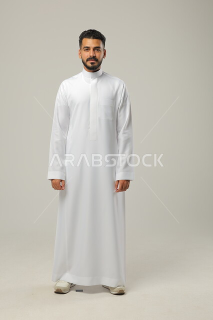 الاهتمام بالمظهر الخارجي والاناقة، مفهوم الرجولة والثقة بالنفس، الوقوف باستعداد واستقامة، بورتريه لرجل عربي خليجي سعودي يرتدي الثوب الأبيض ينظر الى الكاميرا بتعابير السرور،  صورة الجسم بالطول الكامل، خلفية رمادية